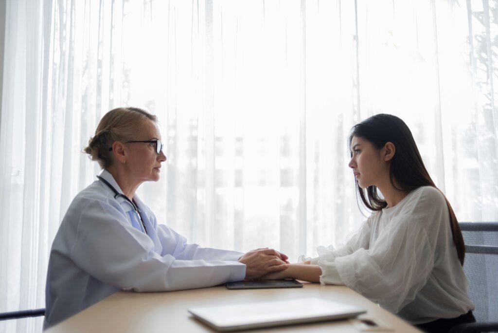 A doctor in a white coat sits across a desk from a young woman, gently holding her hands during a private consultation in a bright office with sheer curtains, both facing each other with serious, attentive expressions as daylight filters through the window behind them.