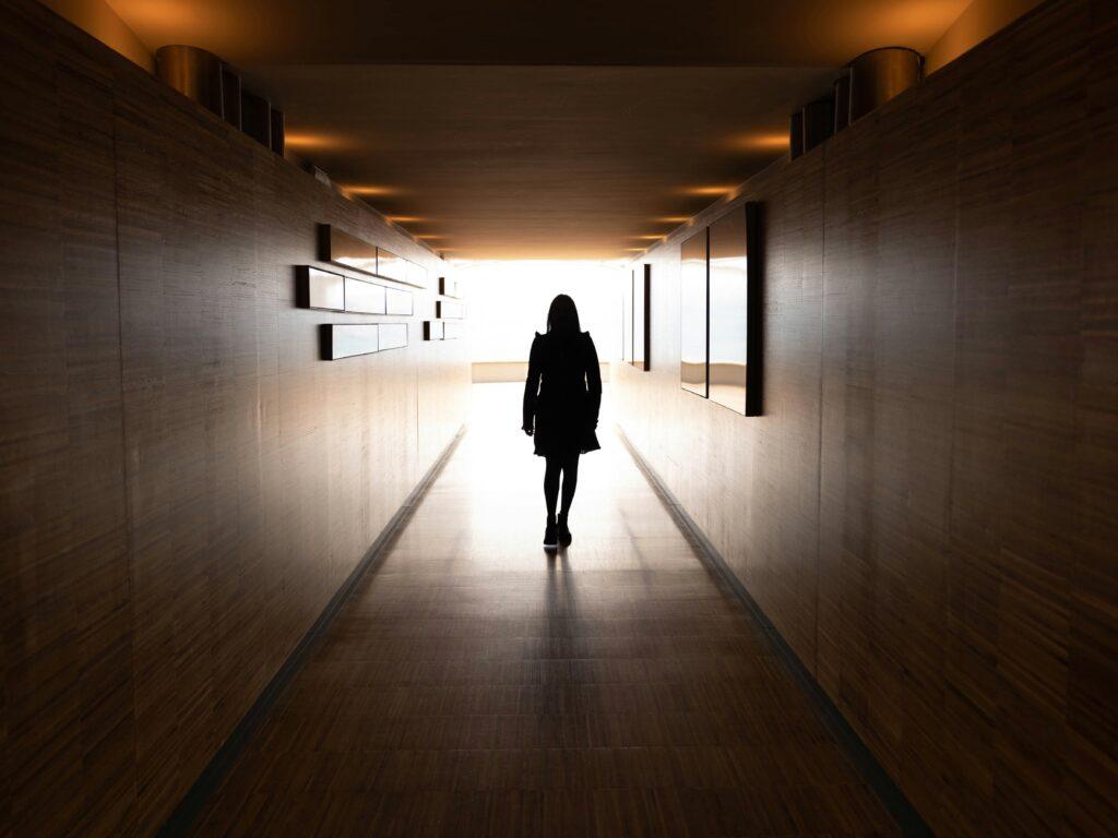 A silhouetted figure walks alone down a long, narrow corridor with wood-paneled walls and warm recessed ceiling lights, moving toward a bright, overexposed opening at the far end that casts strong backlighting and elongated reflections on the polished floor.
