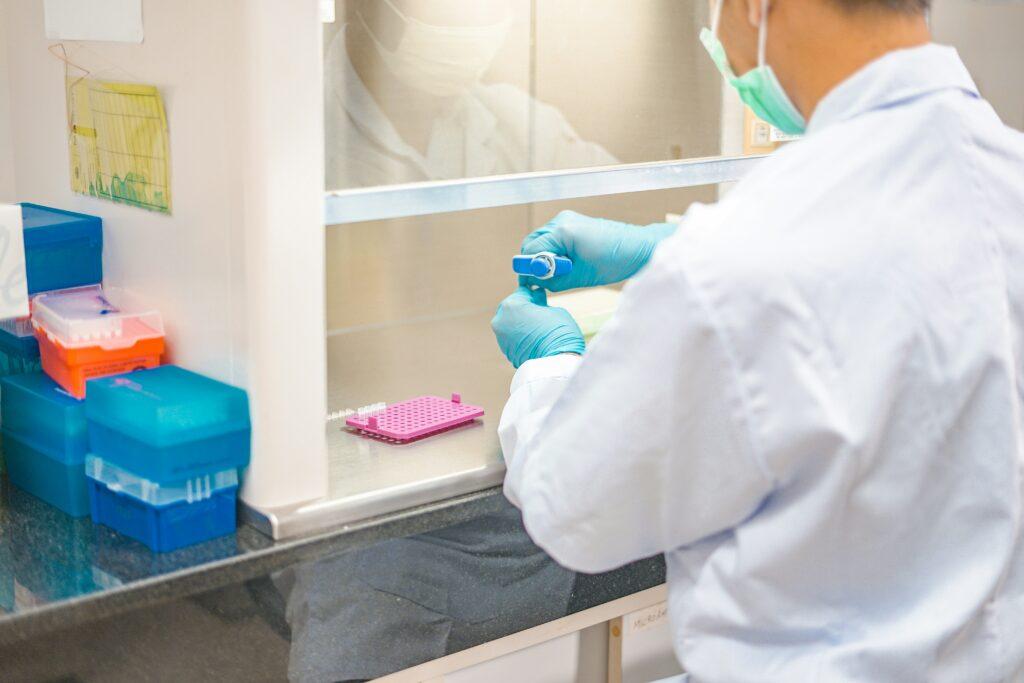 A laboratory technician wearing a white lab coat, face mask, and blue gloves works at a biosafety cabinet, holding a pipette over a small tube, with colorful plastic sample boxes and a pink tube rack arranged on the counter inside the sterile workspace.