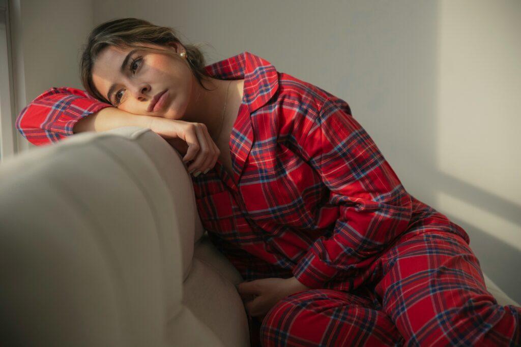 A woman wearing red plaid pajamas reclines on a light-colored couch, resting her head on her forearm against the back cushion while gazing off to the side with a contemplative expression, as warm natural light from a nearby window casts soft shadows across her face and the plain wall behind her.