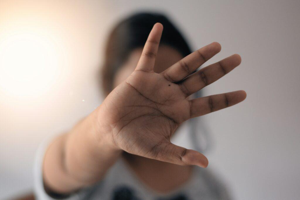 Close-up of an outstretched hand held toward the camera in a clear stopping gesture, palm fully visible with fingers spread, while the person’s face and upper body remain softly blurred in the background against a neutral light-colored wall.