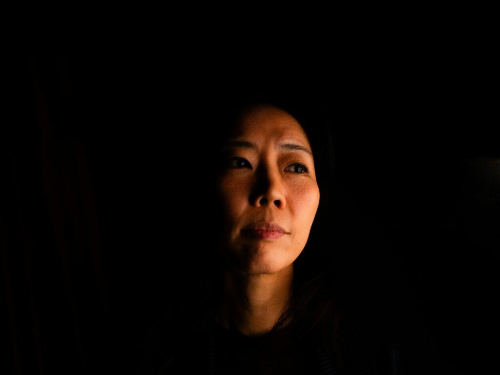 Low-key portrait of a woman emerging from deep shadow, with soft directional light illuminating one side of her face as she gazes off to the side against a dark, nearly black background.