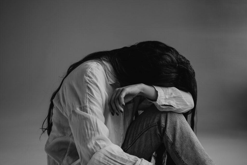 Black-and-white photograph of a person sitting on the floor with knees drawn up, head bowed and partially hidden by their arm, long dark hair falling forward as they wear a light button-up shirt and jeans against a plain, softly lit background.